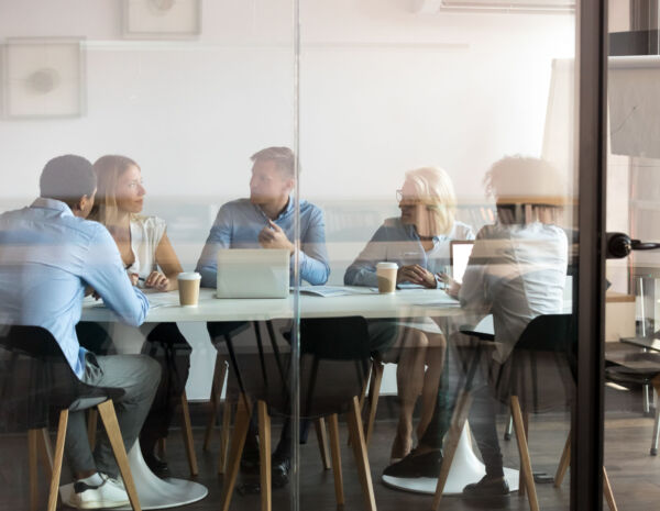 Een blik door het glas op vijf professionals die samenwerken aan een tafel, apparaten gebruiken en het gemak van afdrukken in de cloud laten zien voor naadloze toegang tot documenten.