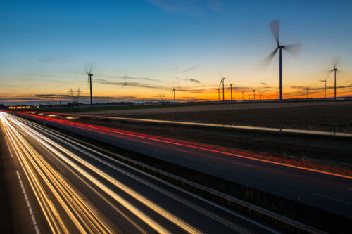 A highway stretches into the distance, flanked by large wind turbines rotating in the breeze during a sunset.