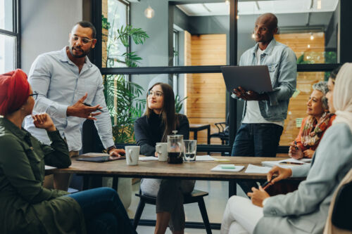 A team of six people in a meeting room discusses reducing IT costs through cloud printing solutions.