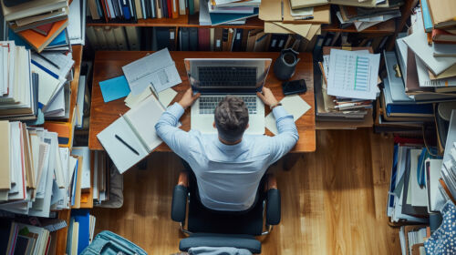 An overhead view at a manl working in a messy workspace with piles of documents, illustrating how cloud printing can reduce print wastage.