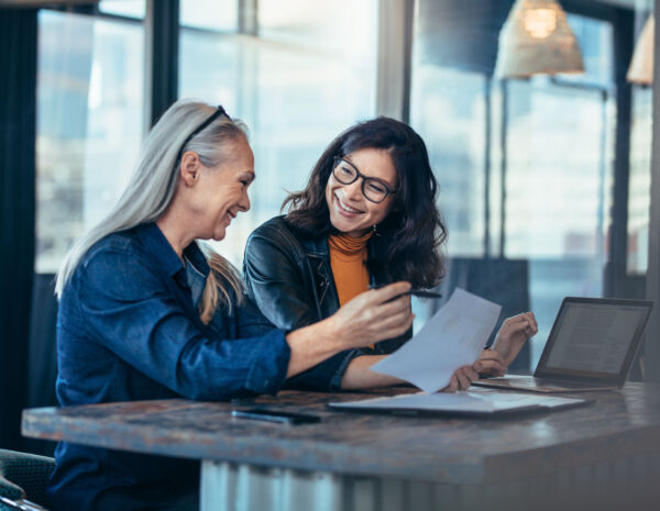 Two smiling businesswomen collaborate around a table in a contemporary workspace, reflecting on how to improve print management.