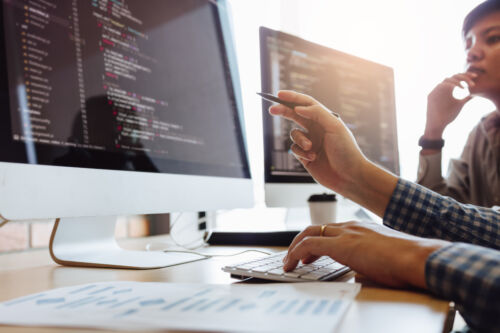 Two professionals work on code at dual computer monitors in an office setting, focusing on a collaborative programming task.
