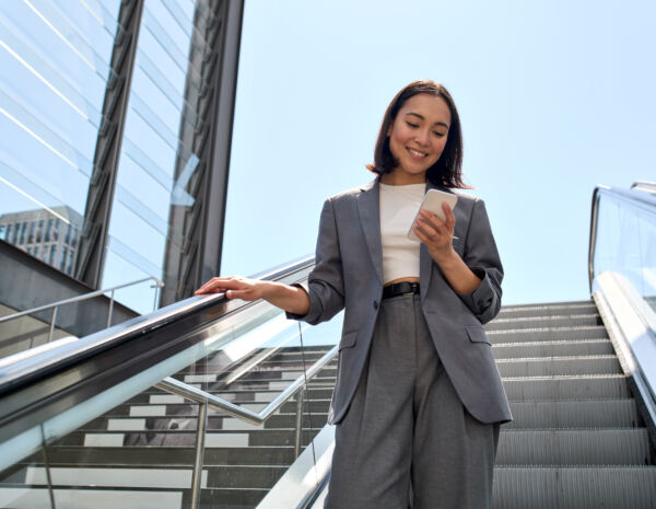 A smiling young female professional in a casual suit stands on an urban escalator, using her smartphone to access the mobile printing.